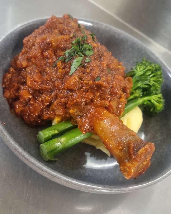 Meat on the bone in rich tomato sauce with mash and green vegetables in a grey bowl